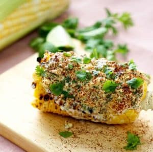 Grilled corn on the cob topped with creamy spice mixture, fresh cilantro, and lime wedge on a wooden board, ideal for relaxing vacation meals in Cozumel Mexico.