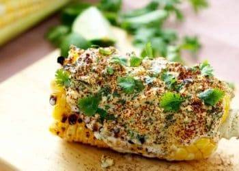 Grilled corn on the cob topped with creamy spice mixture, fresh cilantro, and lime wedge on a wooden board, ideal for relaxing vacation meals in Cozumel Mexico.