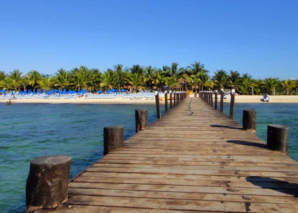 Sunny Cozumel beach with wooden pier leading to turquoise waters, lush palm trees, and blue beach umbrellas, perfect for a relaxing getaway in Mexico.