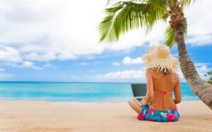 Relaxing woman in a sun hat working on a laptop on the sandy beach in Cozumel, Mexico, under a palm tree, with clear turquoise waters, perfect for vacation rentals and tropical getaways.