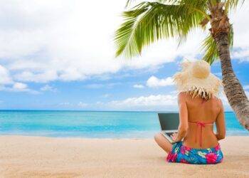Relaxing woman in a sun hat working on a laptop on the sandy beach in Cozumel, Mexico, under a palm tree, with clear turquoise waters, perfect for vacation rentals and tropical getaways.