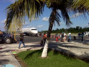 Aircraft on tarmac at Cozumel International Airport with travelers, palm tree in foreground, clear sky, and tropical scenery, offering a preview of the vacation experience near Stingray Villa.