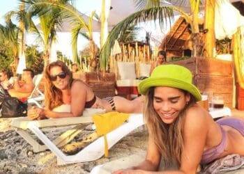 Vibrant Caribbean beach scene at Playa Casitas Beach in Cozumel, Mexico, showcasing relaxed guests sunbathing and enjoying tropical vibes under palm trees with beach loungers and colorful surroundings.