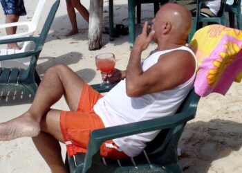 Relaxing man enjoying a cocktail on the beach in Cozumel, Mexico, while smoking a cigar in orange swim trunks.