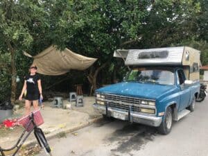 Vintage blue food truck with a camper shell in Cozumel, Mexico, near Stingray Villa vacation rental, with a woman smiling and standing beside a bicycle, surrounded by trees and outdoor seating.