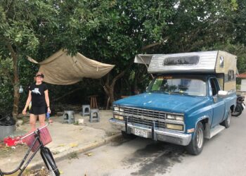 Vintage blue food truck with a camper shell in Cozumel, Mexico, near Stingray Villa vacation rental, with a woman smiling and standing beside a bicycle, surrounded by trees and outdoor seating.