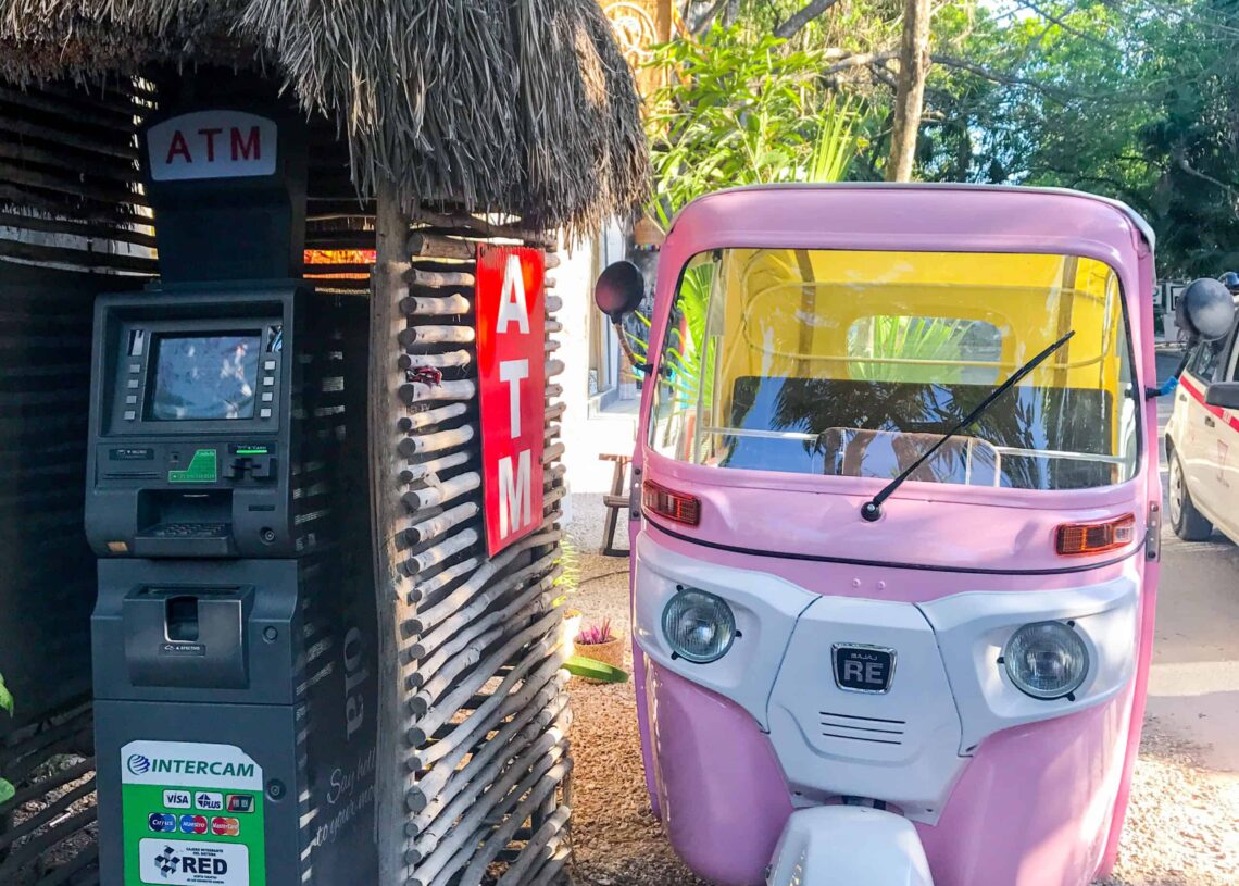 Pink and white auto rickshaw parked next to an ATM machine in a tropical setting.