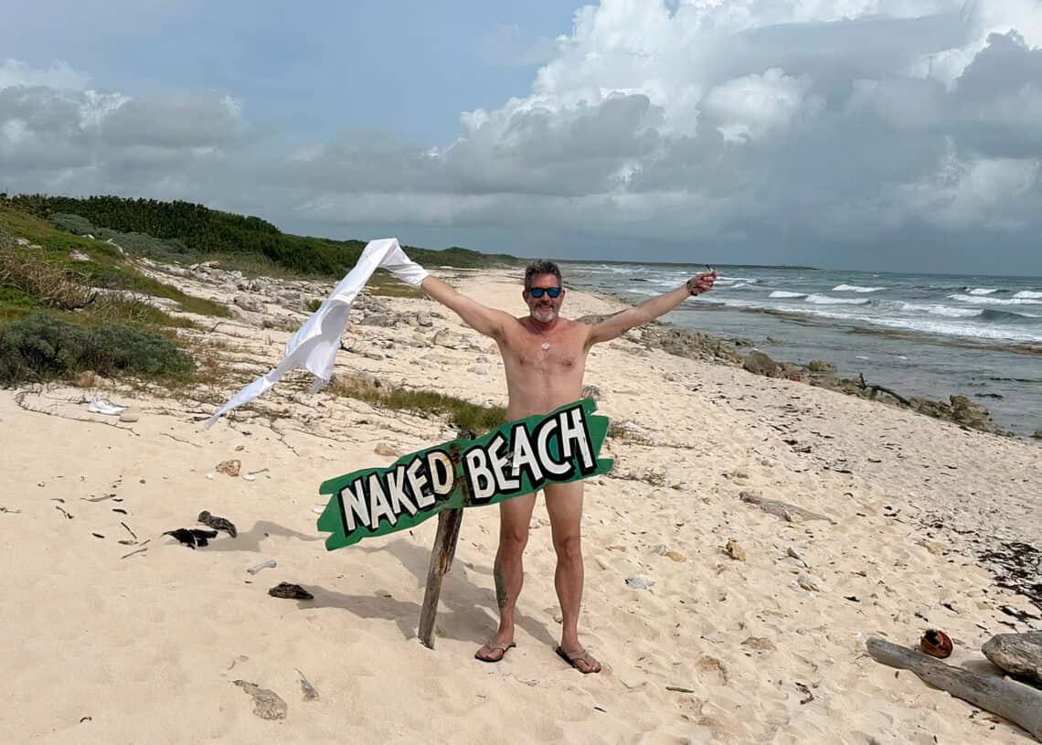 Bright sunny day at a private beach in Cozumel, Mexico, with a man celebrating at Naked Beach sign, surrounded by sandy shores, ocean waves, lush greenery, and a cloudy sky.