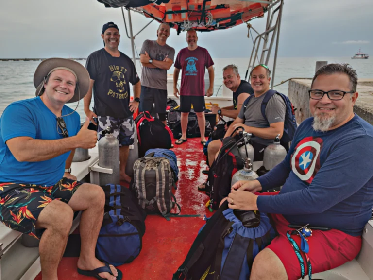 Group of seven smiling Veterans on a boat in Cozumel, Mexico, with luggage and water gear, enjoying a sunny day at sea.