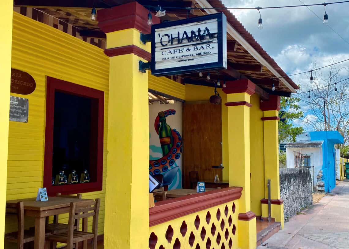 Colorful yellow and red building exterior of Ohana Cafe & Bar in Cozumel, Mexico, with mural art, outdoor seating, and string lights, ideal for vacationers seeking Caribbean island dining.